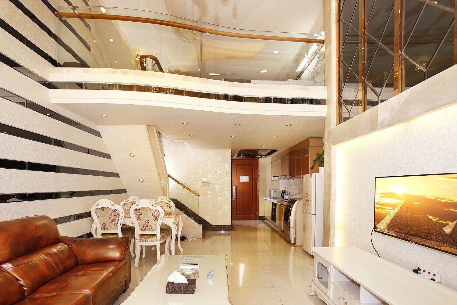 A spacious living area with a high ceiling, a staircase, a kitchen, and a large TV screen displaying a sunset.