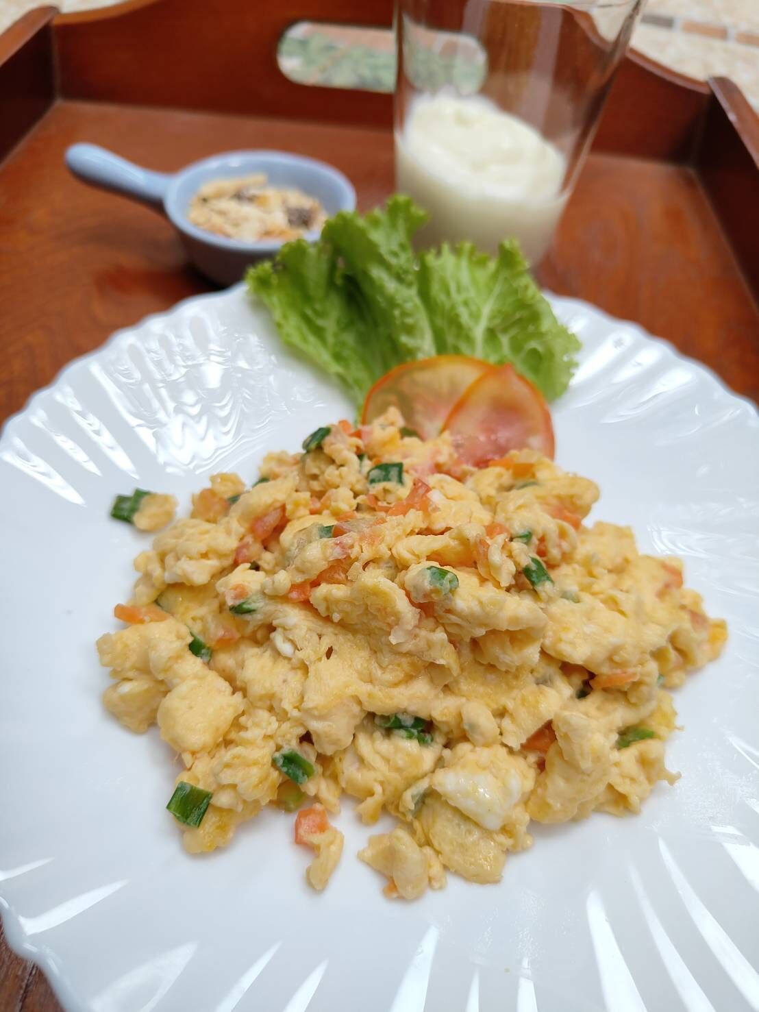 A plate of scrambled eggs with vegetables, a glass of milk, and a small bowl of condiments on a wooden table.