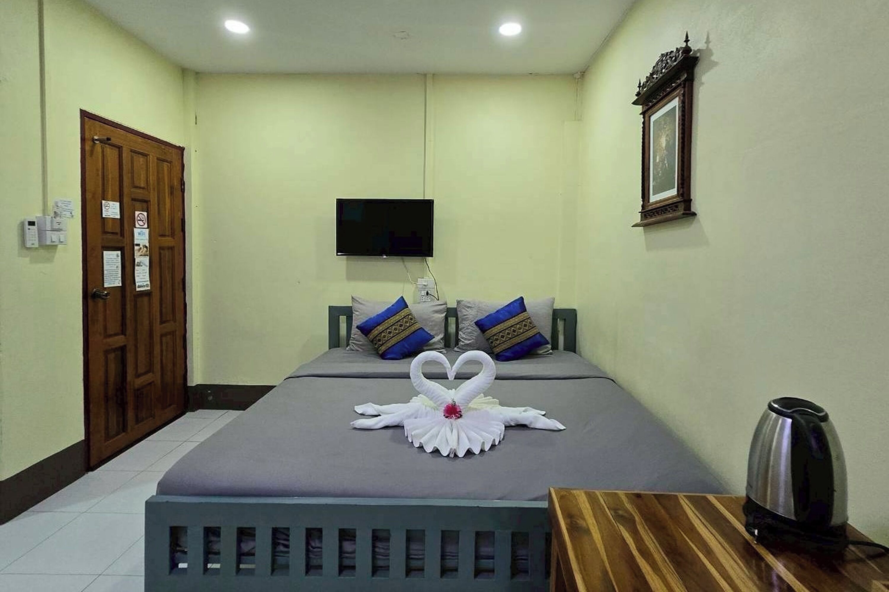 A hotel room with a bed, a television mounted on the wall, a wooden door, and a small table with a kettle.