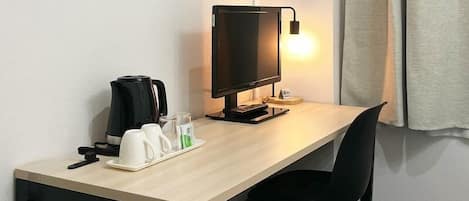 A hotel room with a wooden desk, a black chair, a black monitor, a coffee maker, and a bedside lamp.