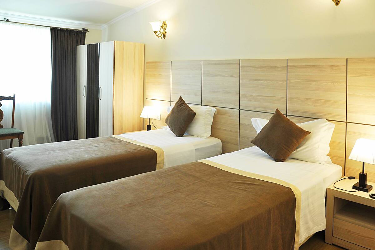 Habitación de hotel con dos camas, cabecera de madera, mesita de noche con lámpara y ventana con cortinas.