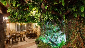 A restaurant with wooden tables and chairs, stone walls, and a rock wall with green foliage. - Hotel Gremi (Tbilisi)