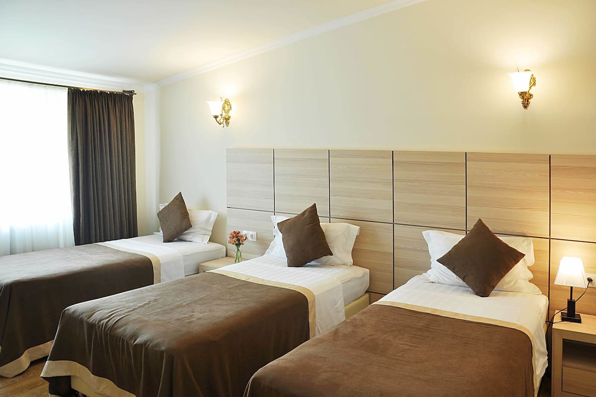 Habitación de hotel con tres camas individuales, cada una con cubrecama marrón y ropa de cama blanca. Hay dos lámparas de pared y una mesita de noche con lámpara.