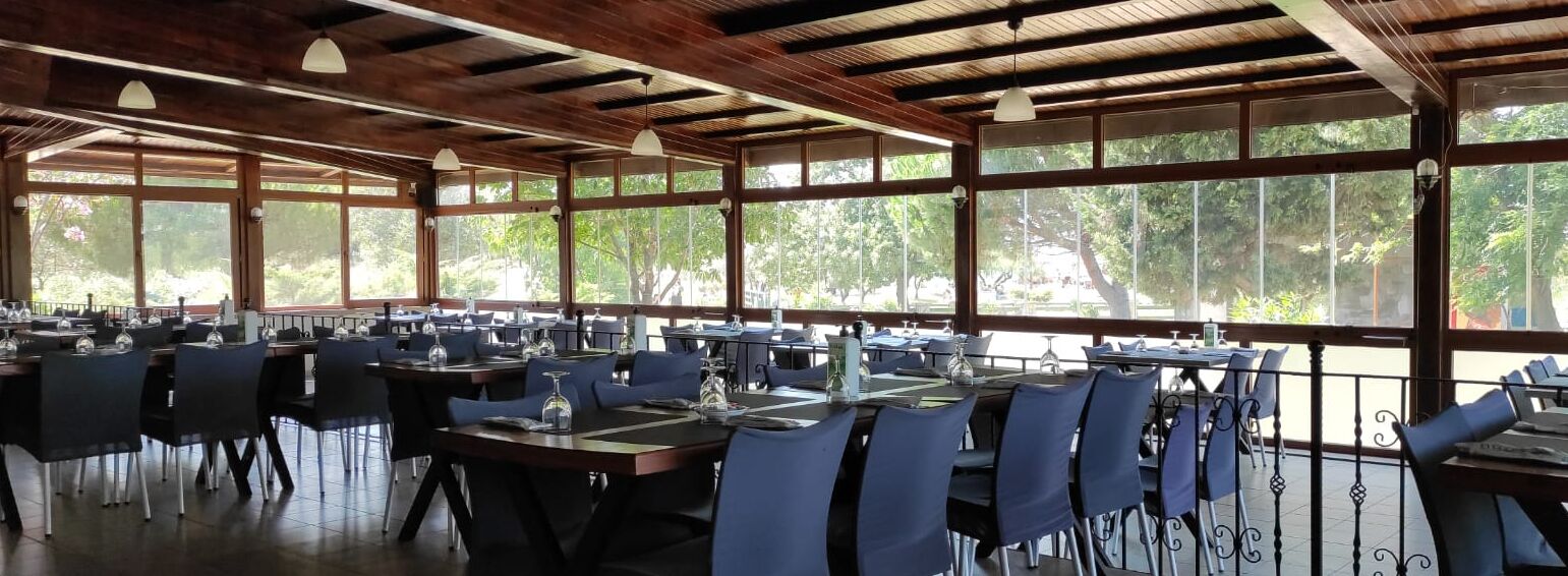 A restaurant with multiple tables and chairs, large windows, and a wooden ceiling.