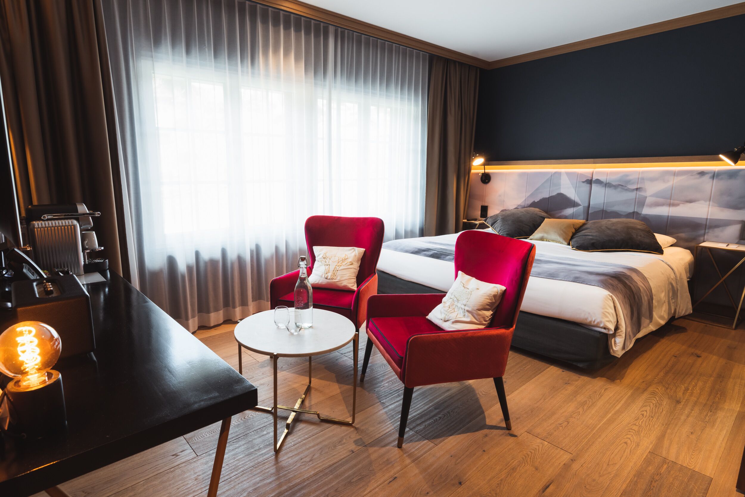 Een hotelkamer met een bed, twee rode fauteuils, een kleine ronde tafel, een bureau met een lamp en een radiator.
