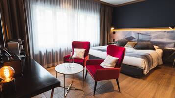 Een hotelkamer met een bed, twee rode fauteuils, een kleine ronde tafel, een bureau met een lamp en een radiator.