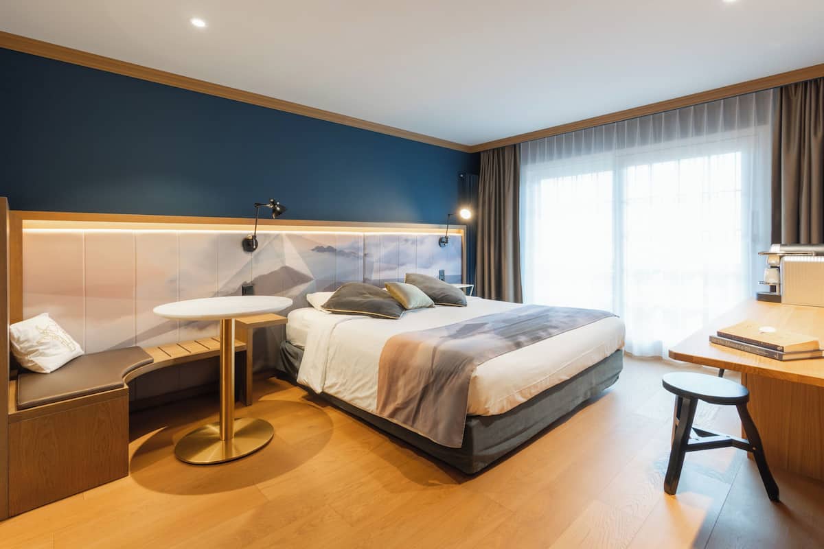 A modern hotel room with a bed, bedside table, round table, and a desk with a chair.