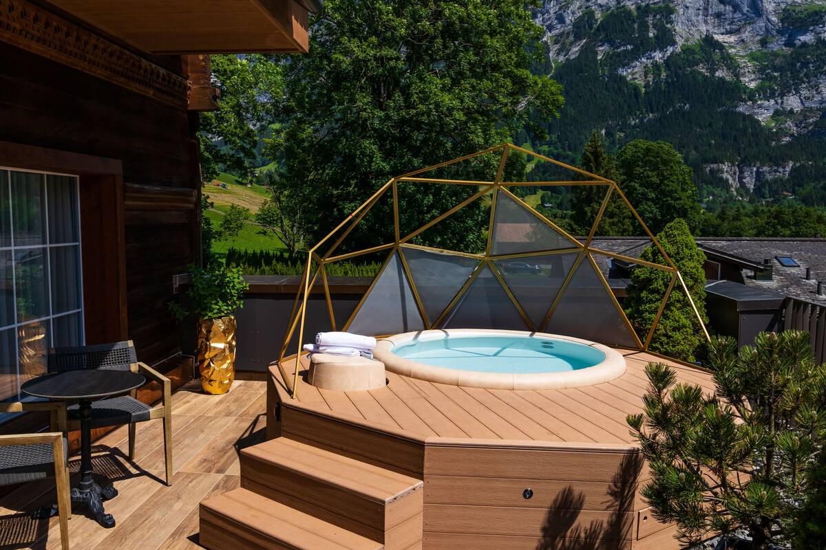 A wooden deck with a hot tub, a glass-enclosed seating area, and a view of a mountainous landscape.
