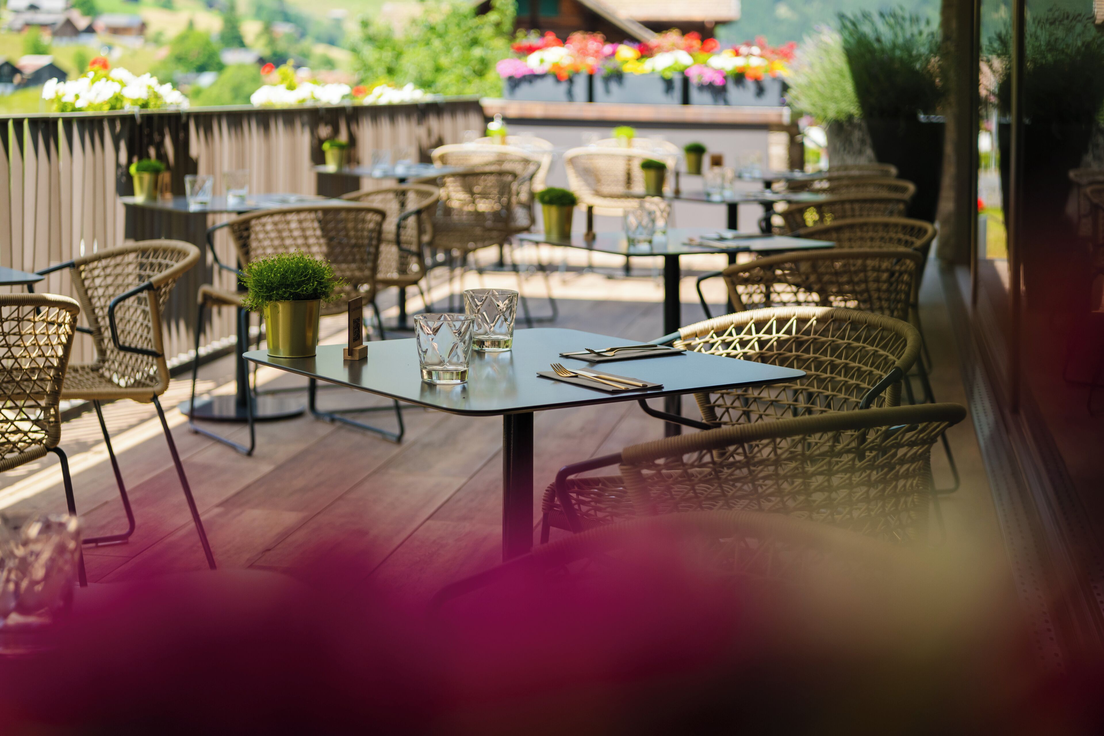 An outdoor dining area with wicker chairs and tables, potted plants, and a view of a garden.