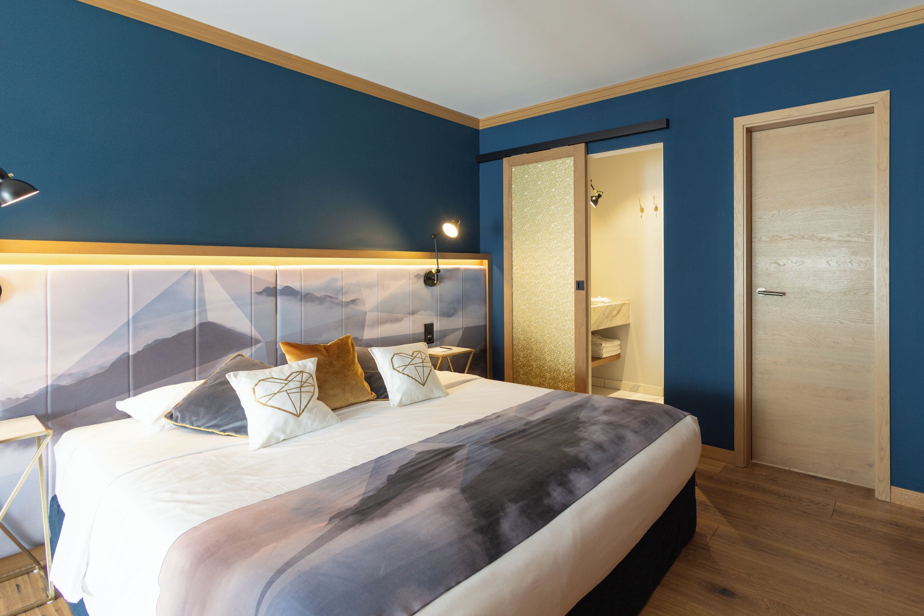 A modern hotel room with a large bed, a nightstand, a desk, a sliding door, and a view of mountains through the window.