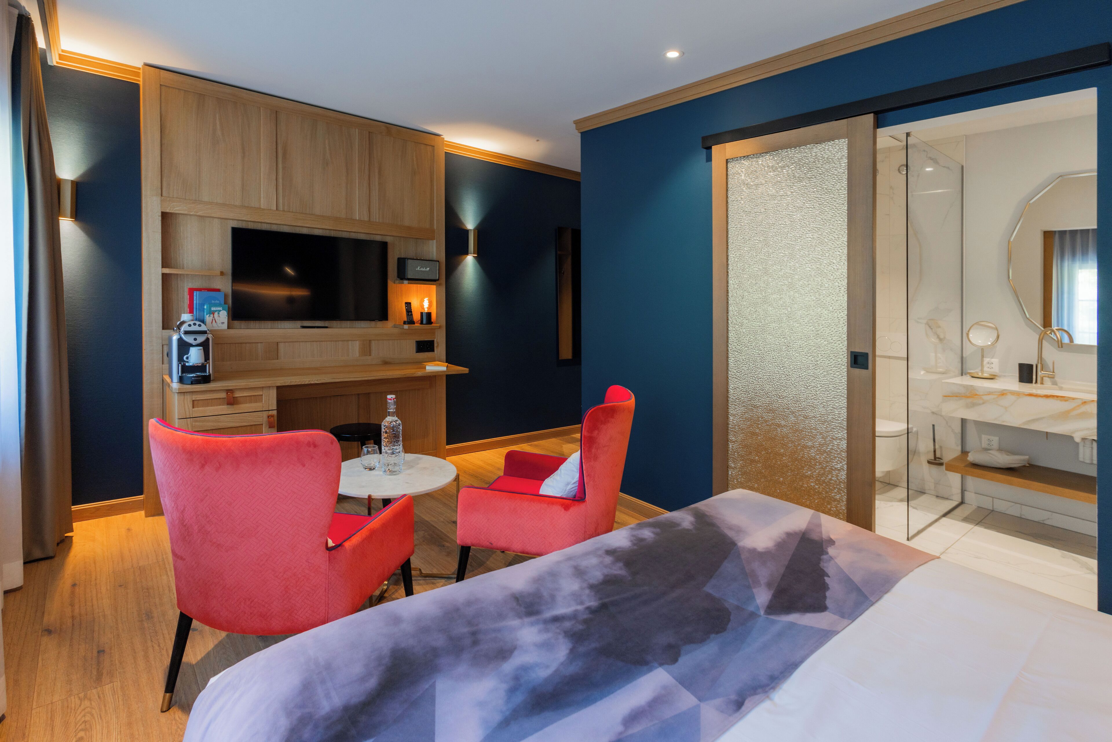 Habitación de hotel con cama, televisión, una pequeña mesa con sillas, minibar y baño con espejo y lavamanos.