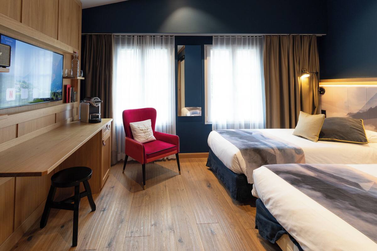 A hotel room with a wooden desk, a red armchair, a bed with a mountain view, and a large window with curtains.