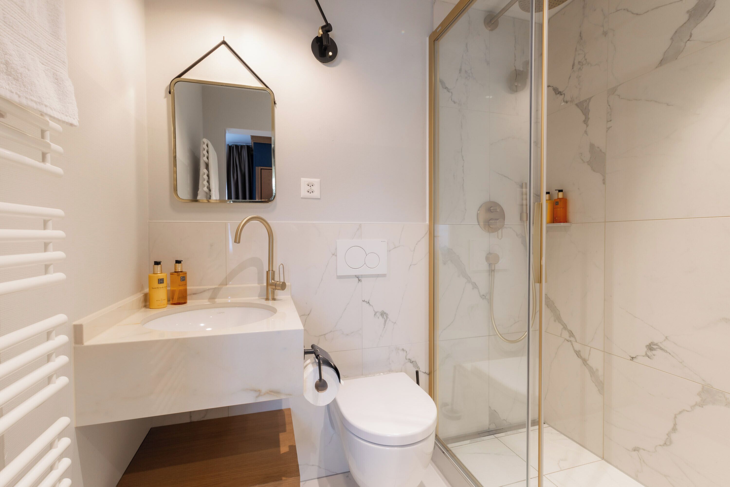 Ein modernes Badezimmer mit einem weißen Waschbecken, einer Toilette, einer Duschkabine aus Glas und einem Spiegel mit goldener Rahmung.