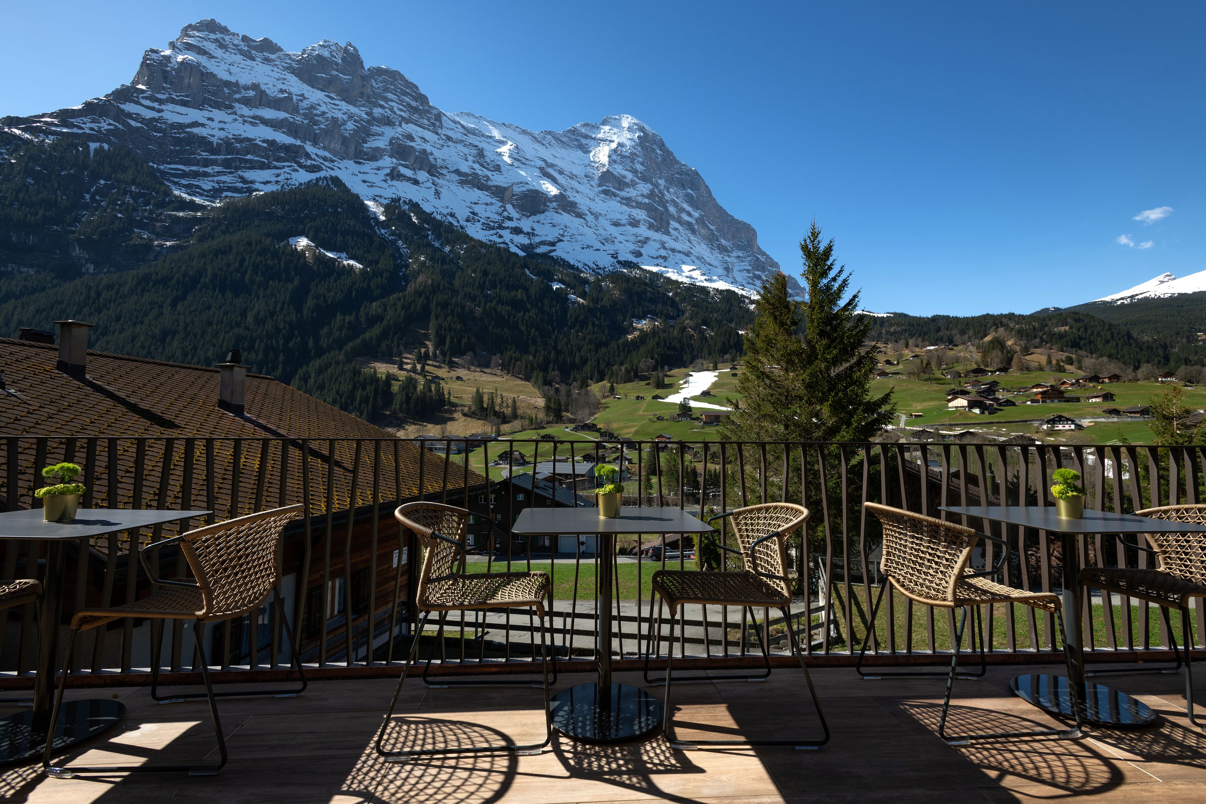 A terrace with wicker chairs and tables, overlooking a mountainous landscape with snow-capped peaks.