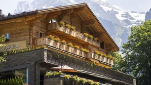 Un chalet en bois doté de balcons ornés de fleurs, une enseigne indiquant «Glacier», et une chaîne de montagnes en arrière-plan.