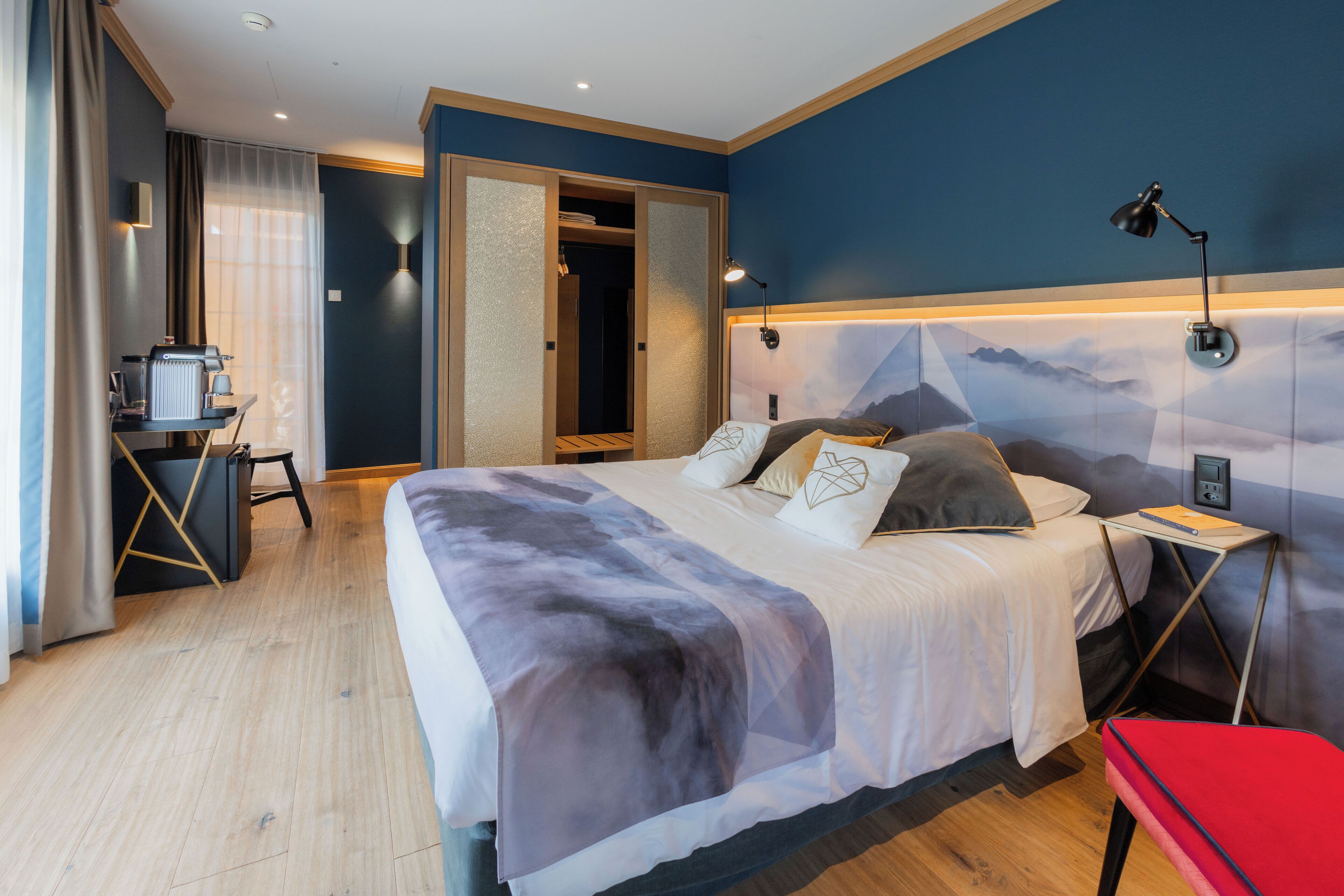 Ein modernes Hotelzimmer mit einem großen Bett, einem Schreibtisch mit Kaffeemaschine, einem roten Sessel und Blick auf die Berge durch das Fenster.