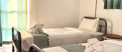 A hotel room with two single beds, a bedside table with a lamp, an air conditioning unit, and a window with curtains.