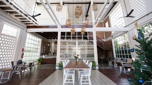 Un espacio interior amplio con una larga mesa de madera rodeada de sillas blancas, un árbol de Navidad y un balcón con barandilla.