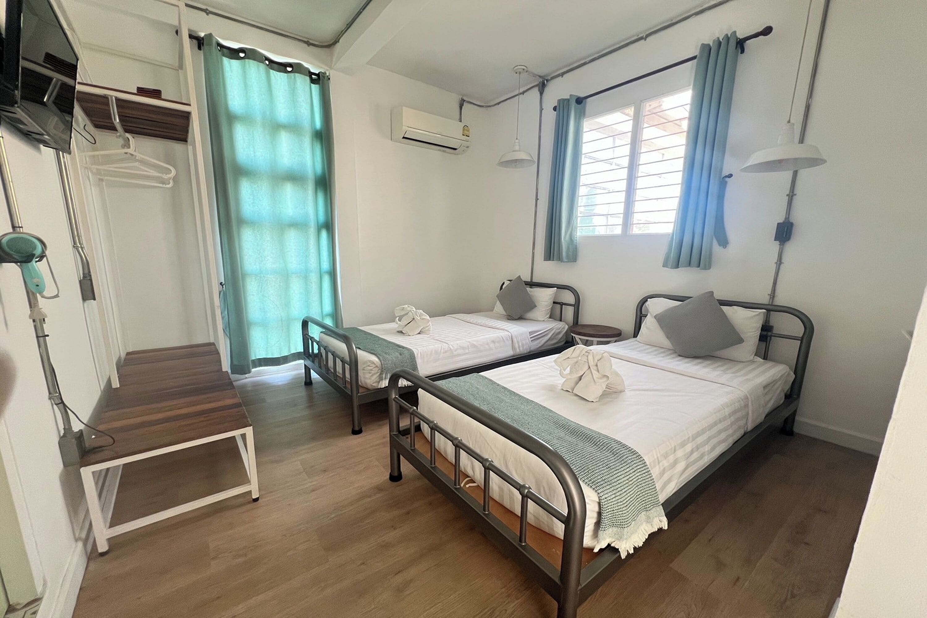 A hotel room with two single beds, a small table, a bench, and a window with curtains.