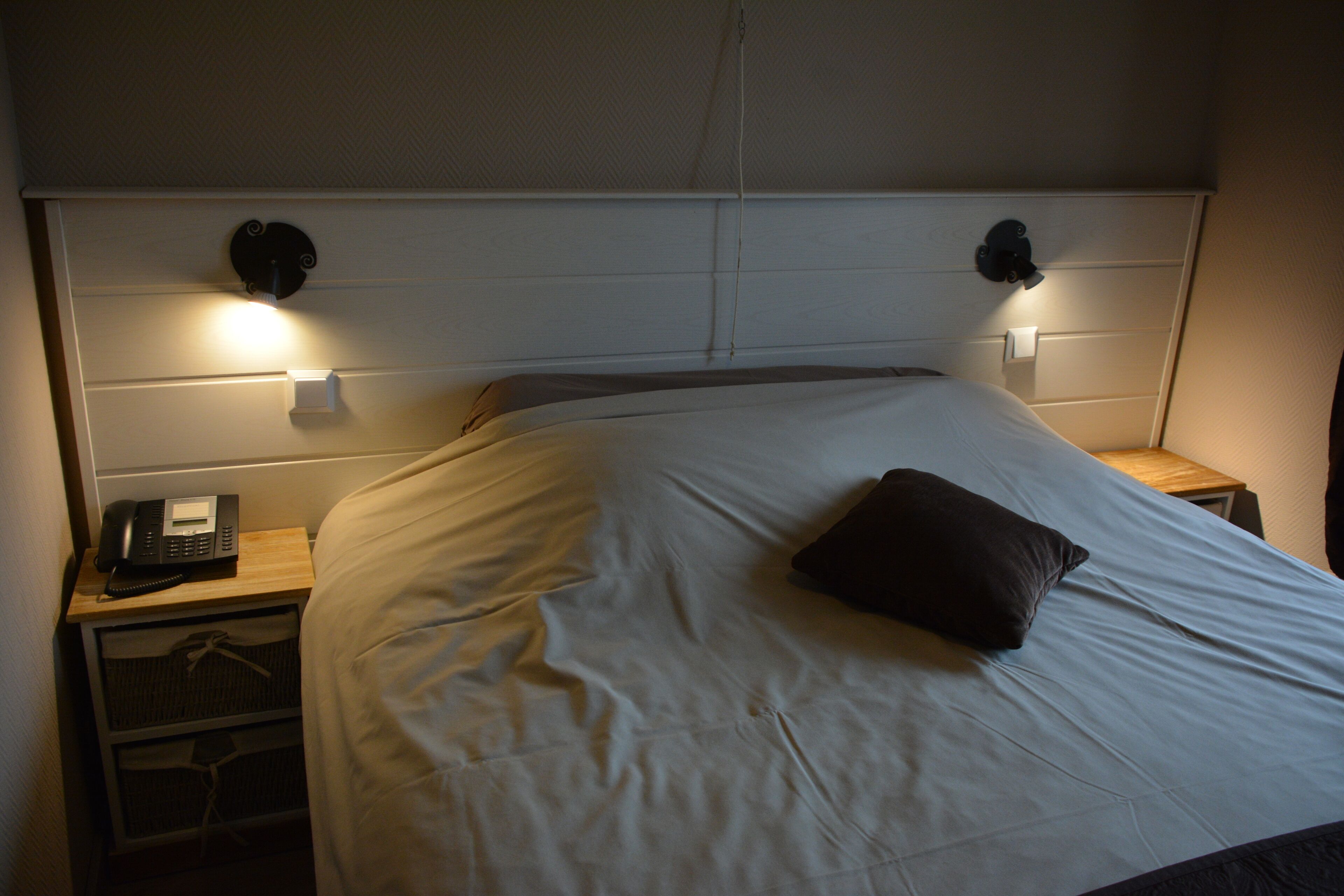 Una cama bien hecha con un cojín, un teléfono en la mesita de noche y dos lámparas de pared a cada lado.
