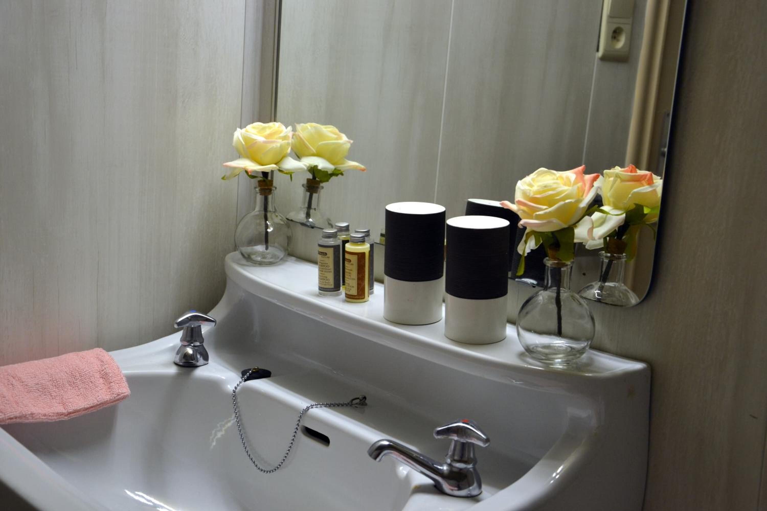 A bathroom sink with two faucets, a pink towel, and three vases with flowers and toiletries.