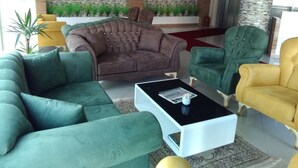 Un salon moderne avec un canapé vert, un fauteuil marron et un fauteuil jaune. Un téléviseur à écran plat est fixé à un mur en pierre, ainsi qu’un élément décoratif mural et une plante en pot.