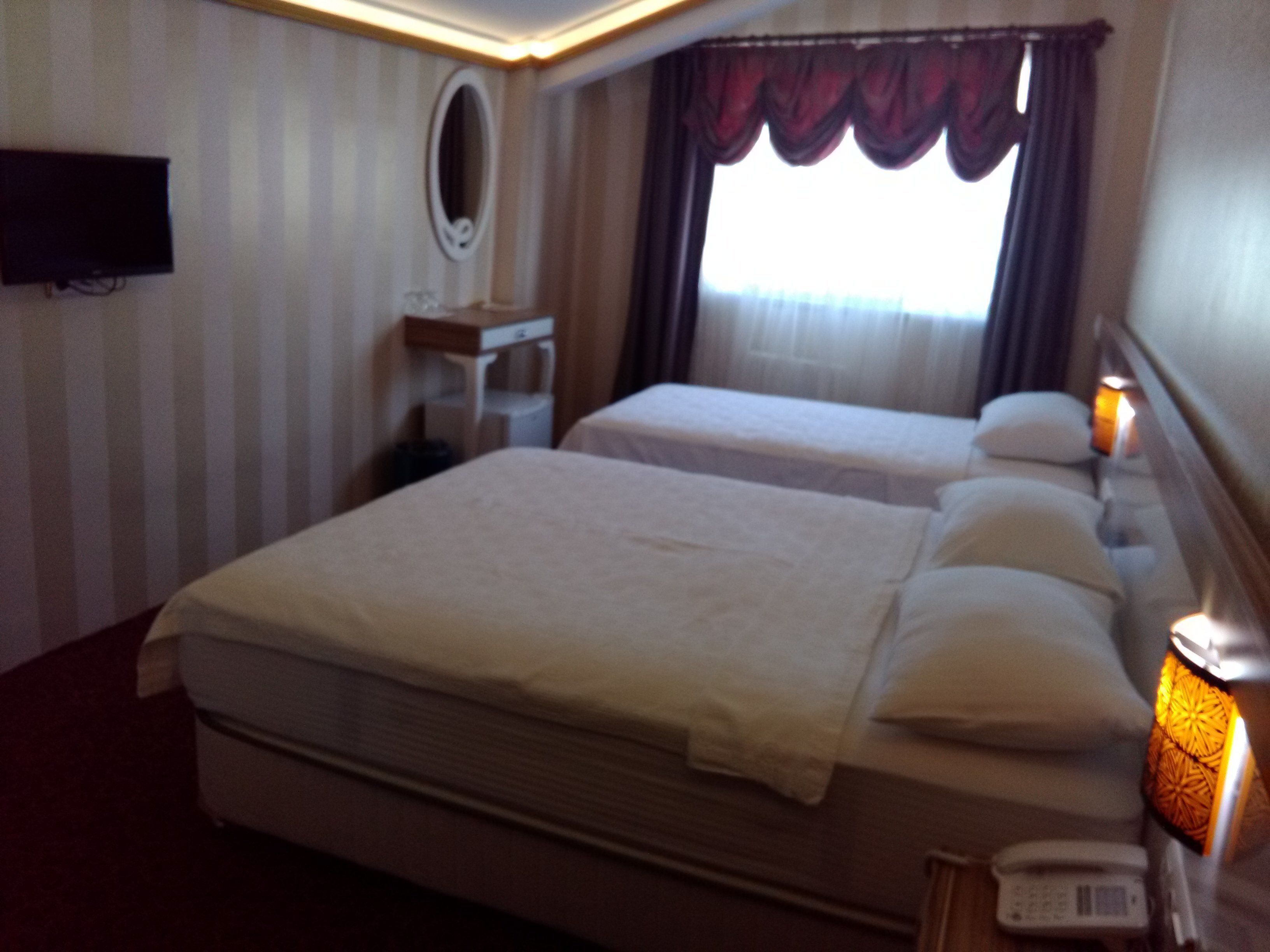 A hotel room with two beds, a bedside table with a lamp, a telephone, and a window with curtains.