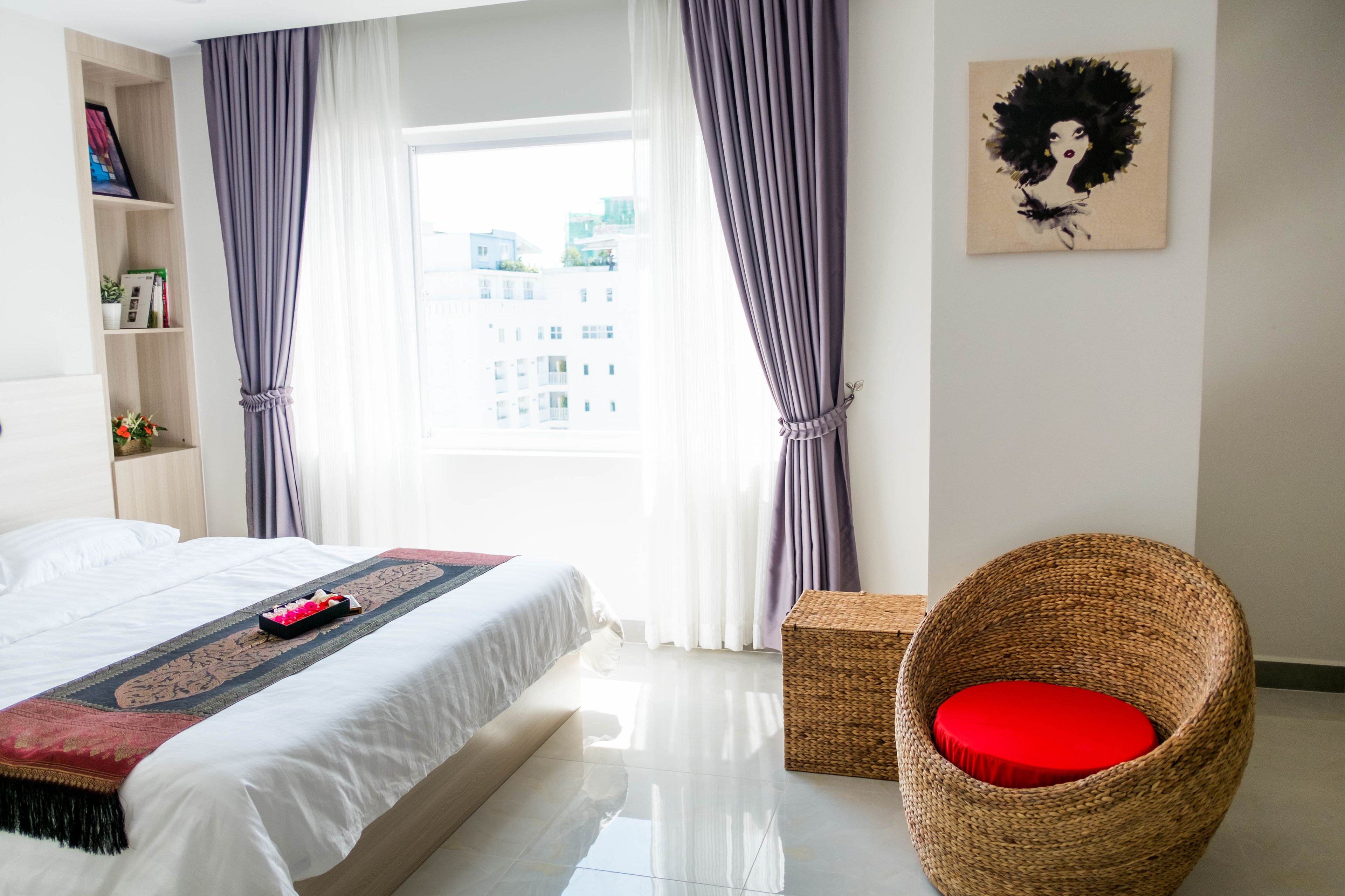 A hotel room with a bed, a wicker chair with a red cushion, a bookshelf, and a painting on the wall.