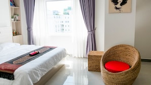 A hotel room with a bed, a wicker chair with a red cushion, a bookshelf, and a painting on the wall. - Service One (Phnom Penh)