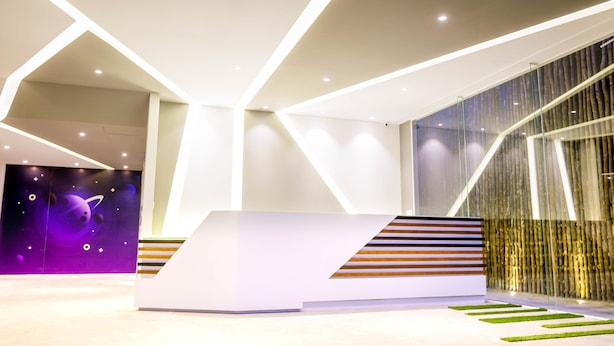 A modern reception area with a white reception desk, geometric ceiling design, and a glass wall with a vertical wooden panel. - Service One (Phnom Penh)