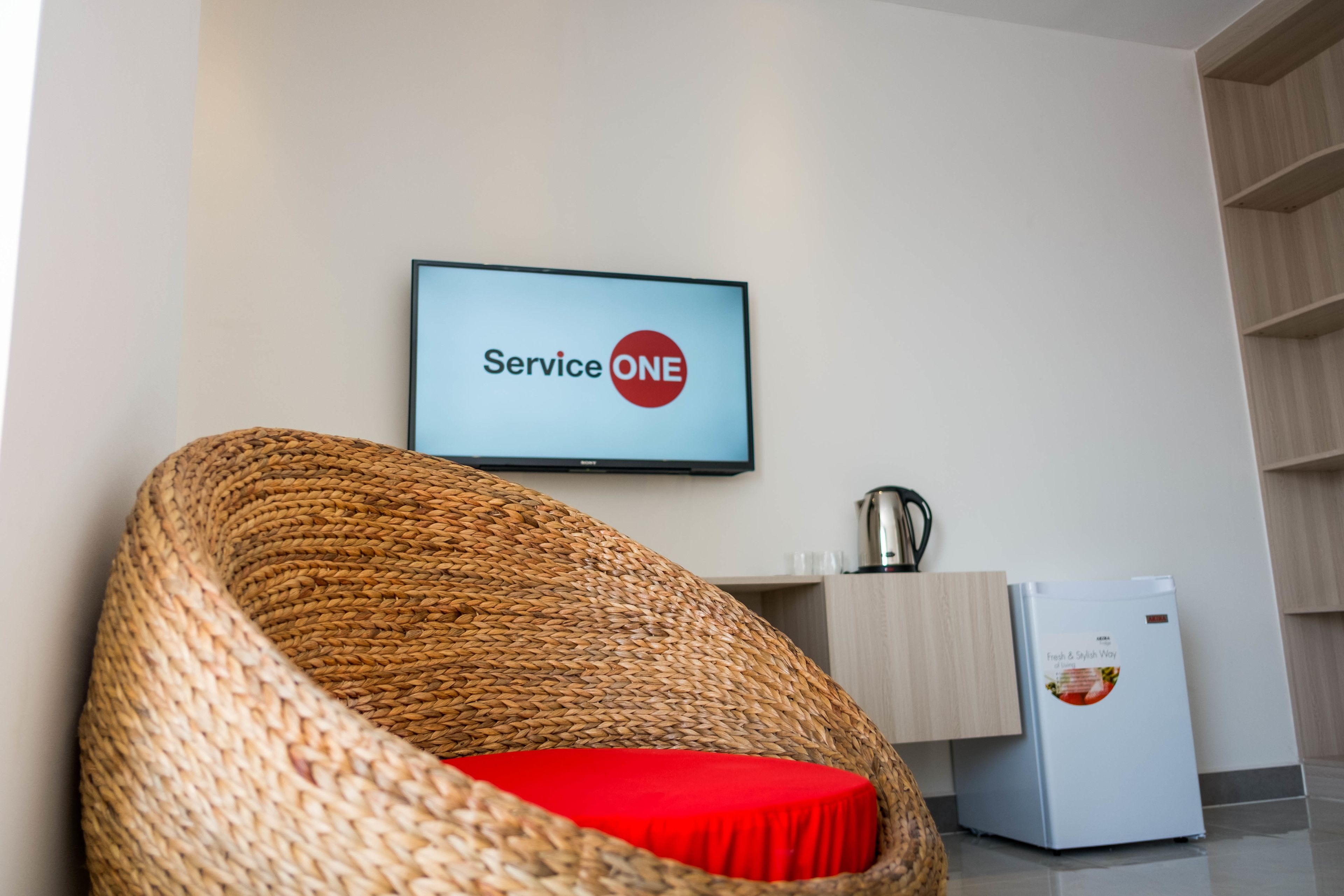 A wicker chair with a red cushion, a television displaying "Service ONE", a small refrigerator, and a wooden cabinet.