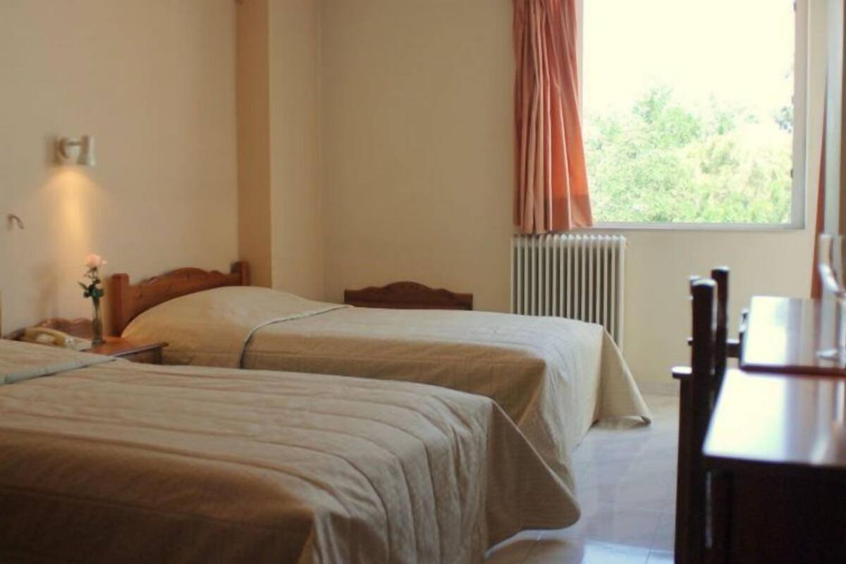 Une chambre d’hôtel avec deux lits, une table de chevet, un vase avec des fleurs, un radiateur et une fenêtre avec des rideaux.