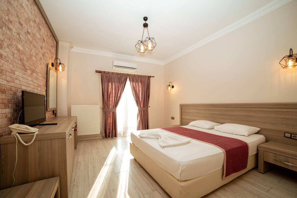 Una habitación de hotel con un escritorio de madera, una cama con una colcha roja, cabecera de madera y una pared de ladrillo.