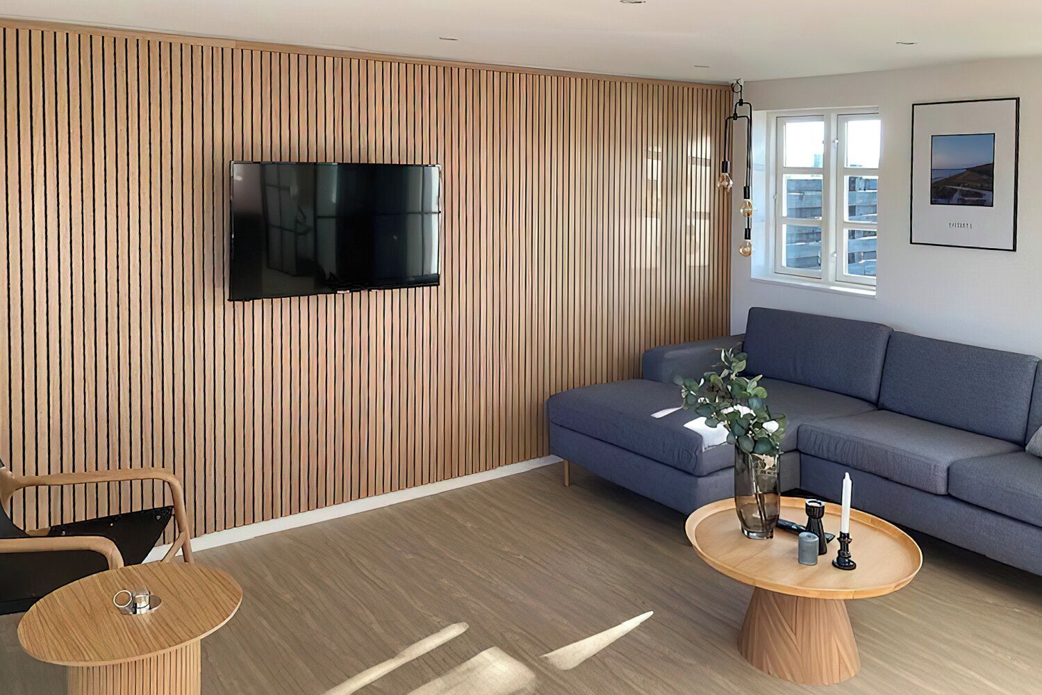 A modern living room with a wooden slat wall, a flat-screen TV, a grey sofa, a round wooden coffee table with decorative items, and a window with a city view.