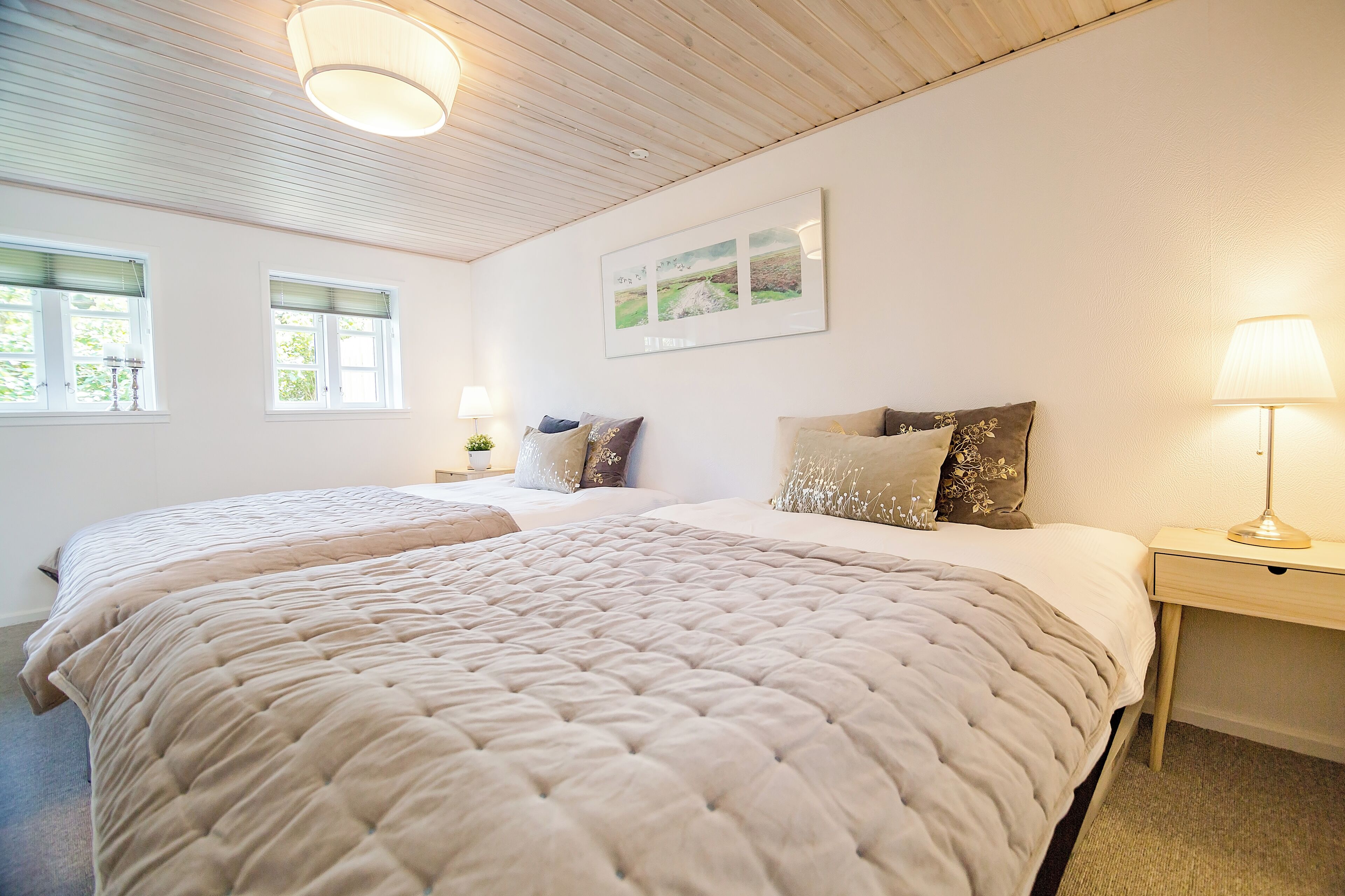 Una cama doble con cubrecama acolchado color beige, dos mesitas de noche con lámparas y un cuadro de paisaje enmarcado en la pared.