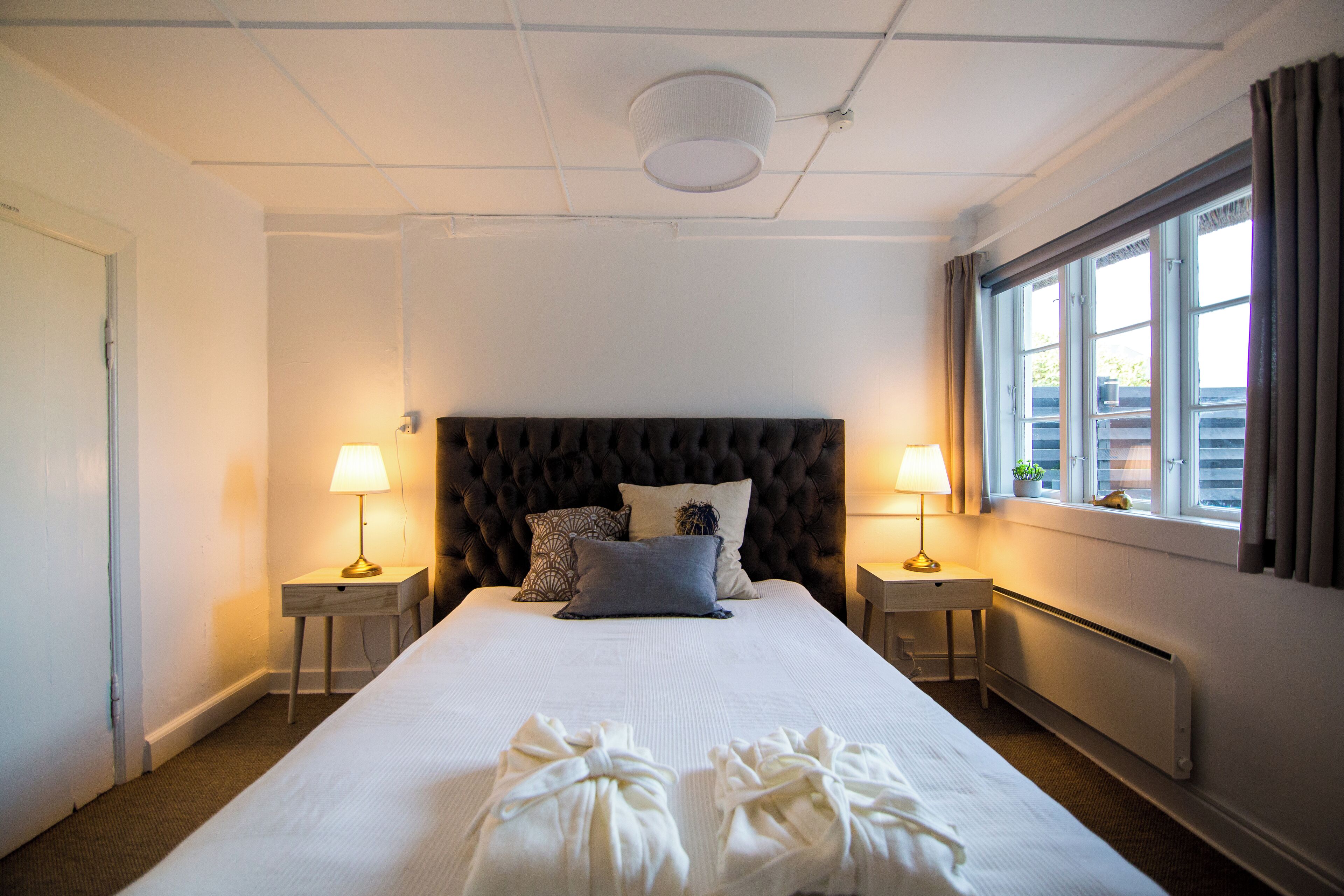 A hotel room with a large bed, two bedside tables with lamps, a window with curtains, and a radiator.