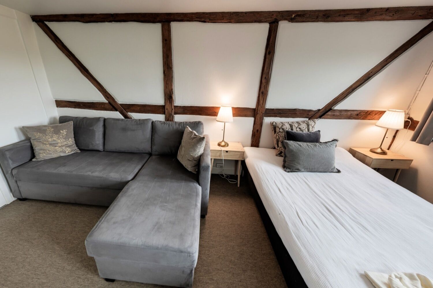 A room with a grey sofa, a single bed, and a nightstand with a lamp.