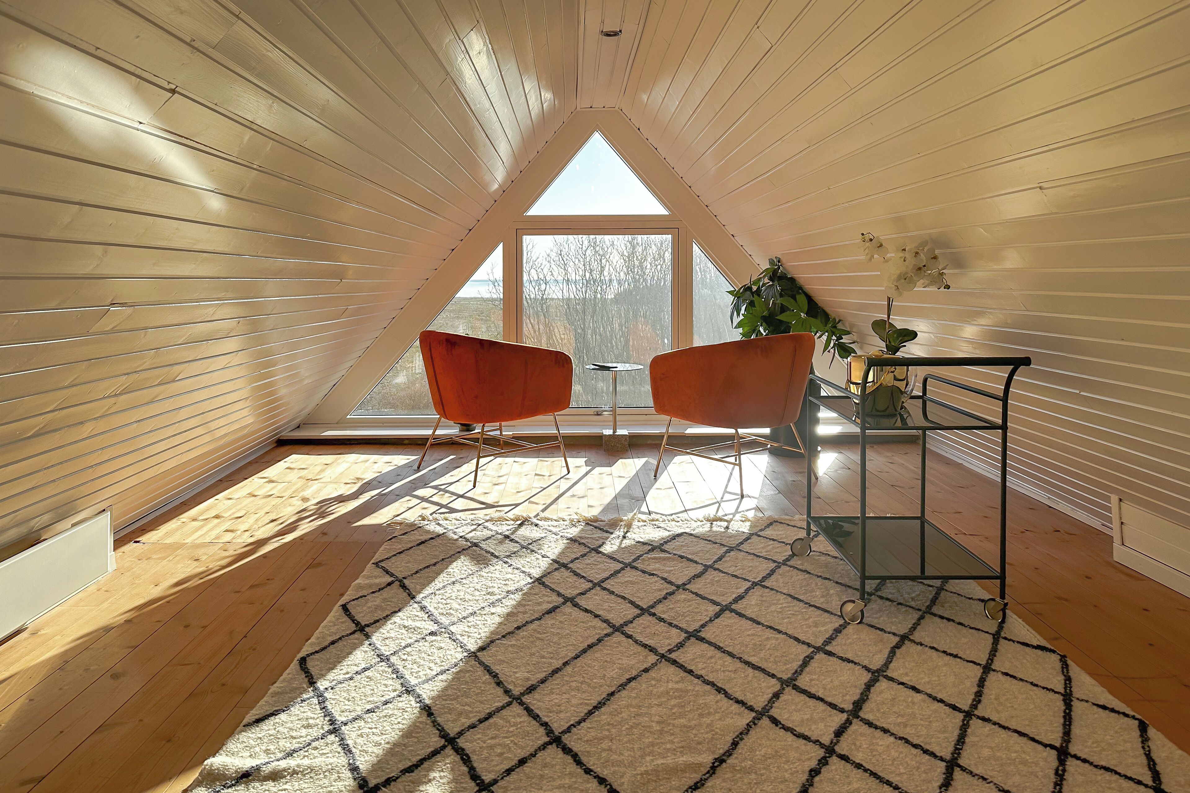 A cozy attic room with wooden walls, a large window, two orange chairs, a small table, and a potted plant.