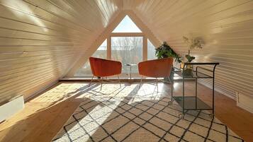 A cozy attic room with wooden walls, a large window, two orange chairs, a small table, and a potted plant.
