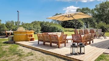 En træterrasse med udendørs møbler, herunder sofaer og et bord, samt en stor parasol der giver skygge.