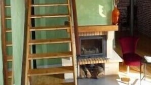 Een rustiek interieur met een houten wenteltrap naar een vide, een open haard en een rode stoel.