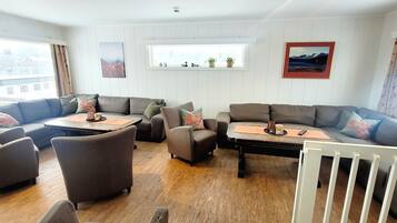 Una sala de estar acogedora con un sofá seccional, sillones y una mesa de centro. Hay dos ventanas con cortinas, una chimenea y vistas al exterior.