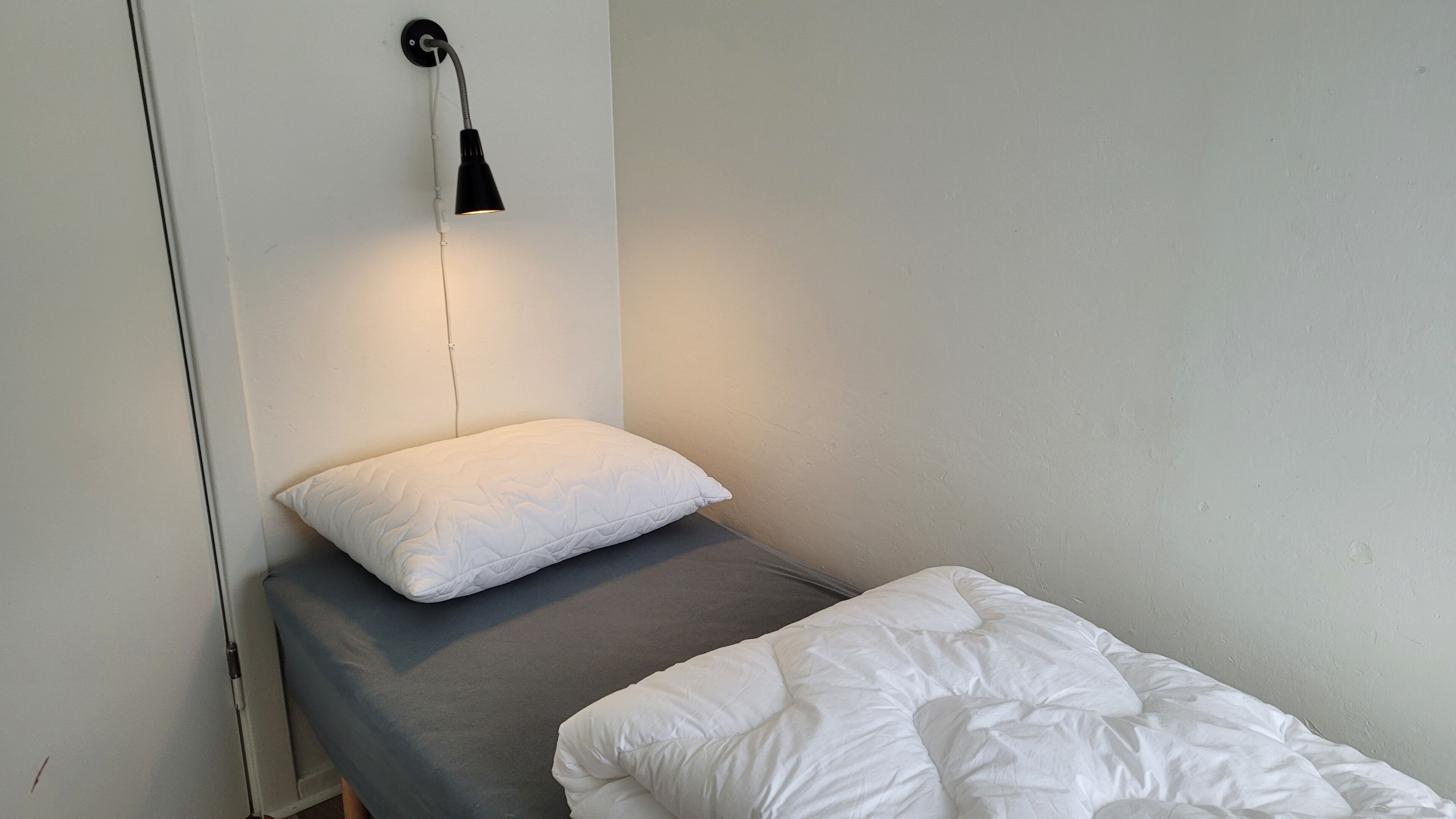 A small, single-bed room with a grey mattress and white bedding. A black wall-mounted lamp provides light, and a white pillow is placed on the bed.