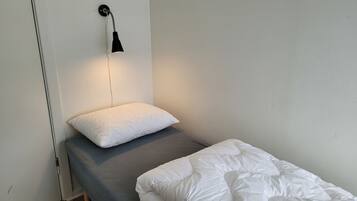 A small, single-bed room with a grey mattress and white bedding. A black wall-mounted lamp provides light, and a white pillow is placed on the bed.