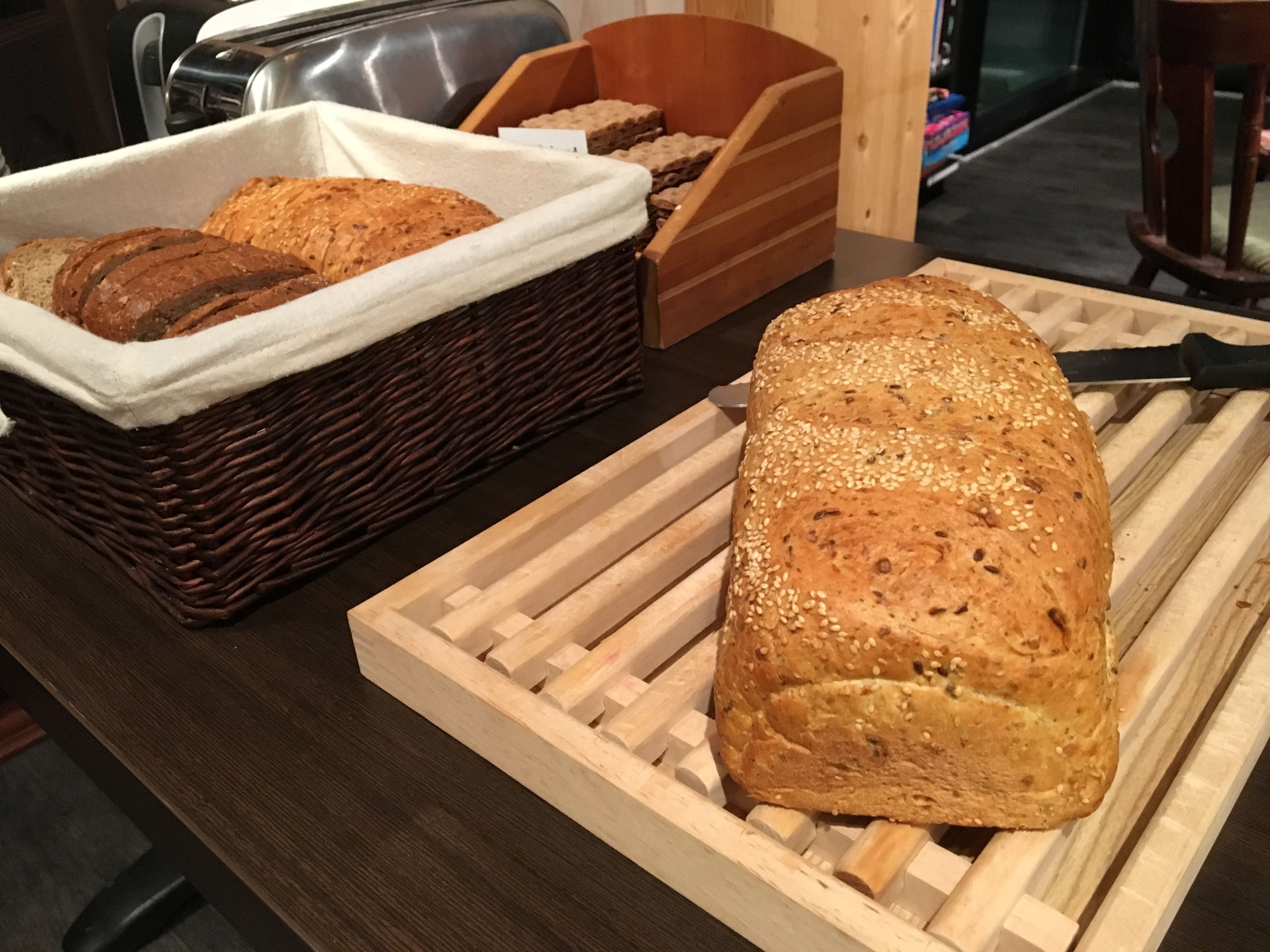 A loaf of bread on a wooden cutting board, a wicker basket with bread, and a toaster in the background.