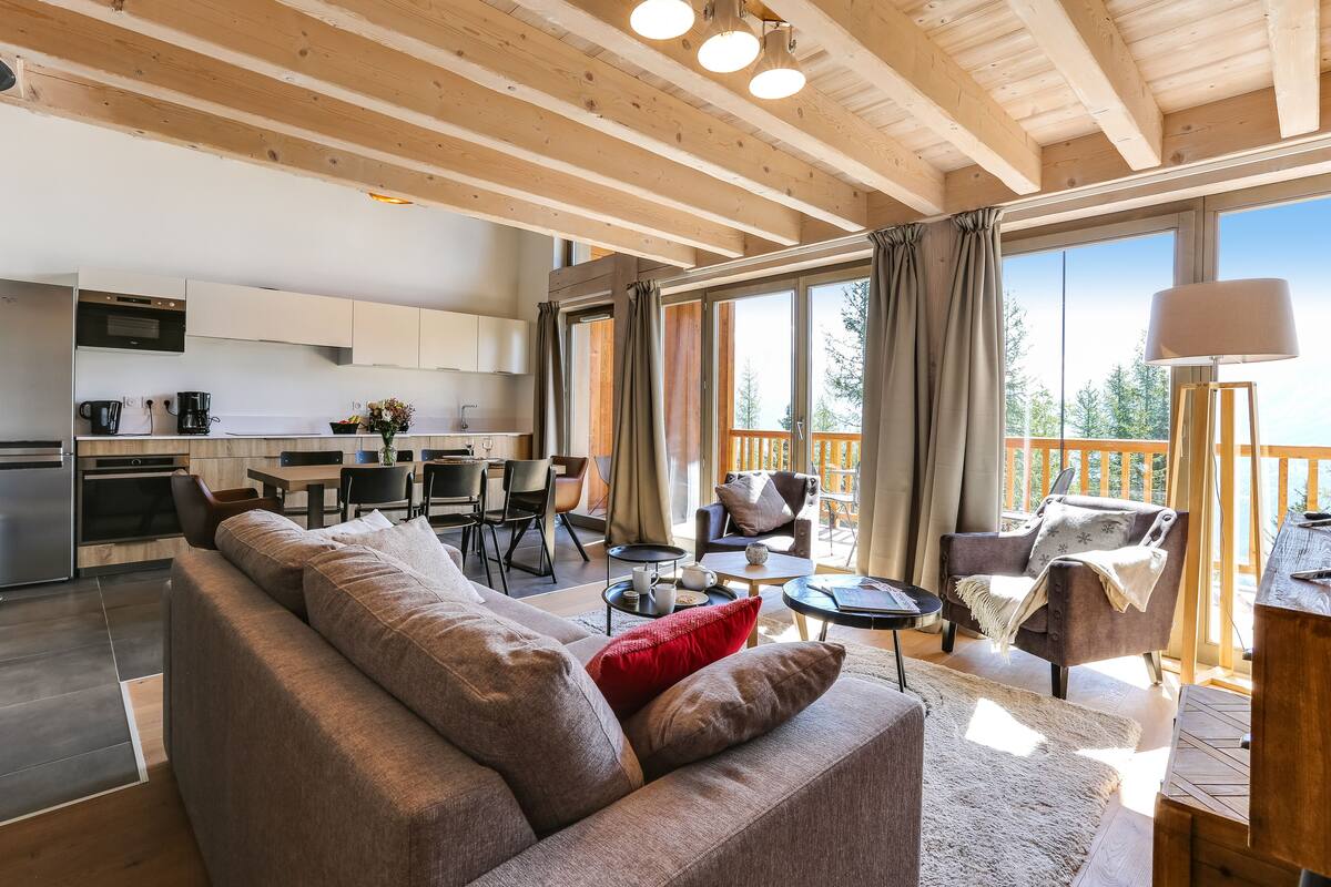 Un salon moderne avec un canapé, une table basse et un coin repas. Au fond, on aperçoit une cuisine avec une table et des chaises. De grandes fenêtres laissent entrer la lumière naturelle et offrent une vue sur l’extérieur.