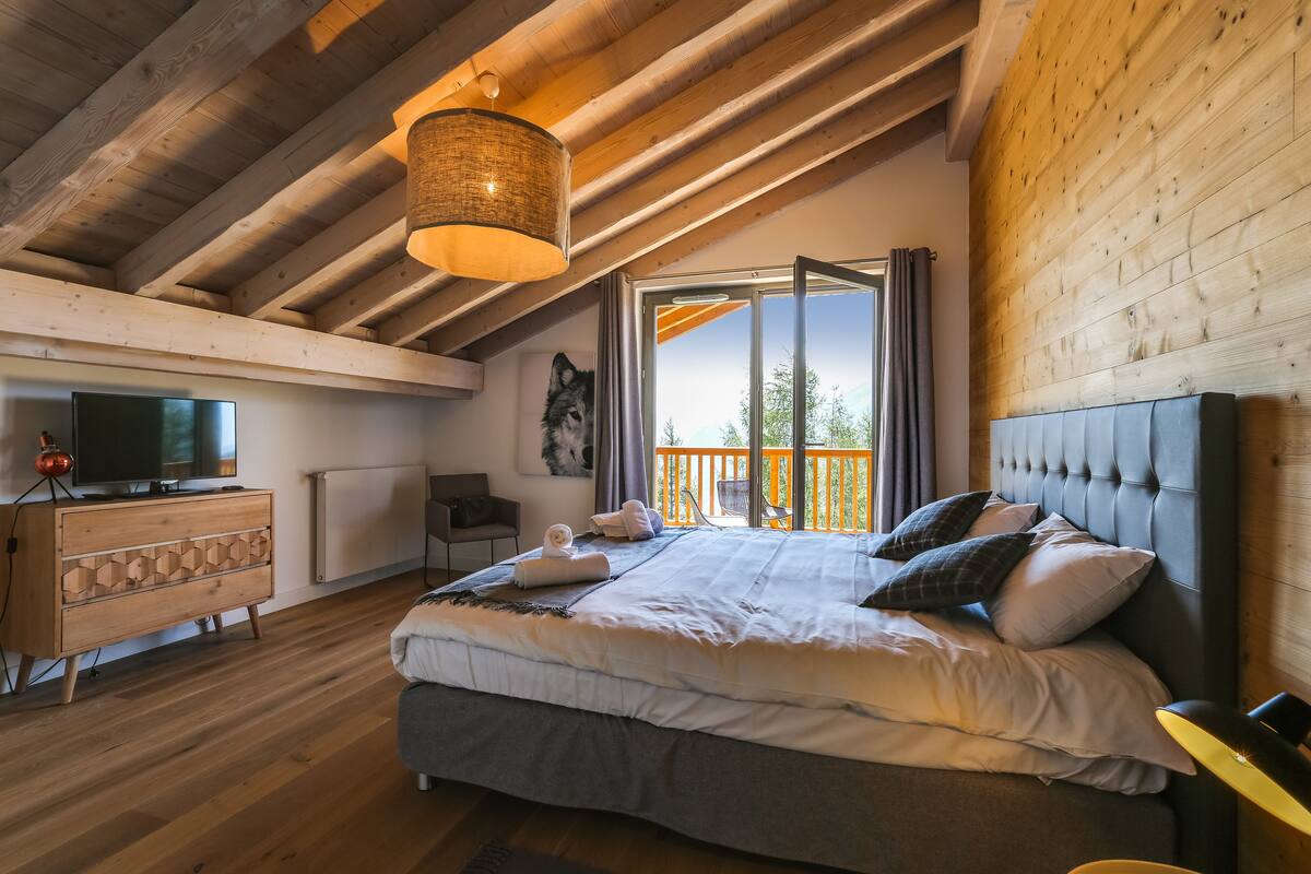 Une chambre dotée d’un grand lit, de poutres apparentes en bois au plafond, d’une télévision et offrant une vue sur l’extérieur.