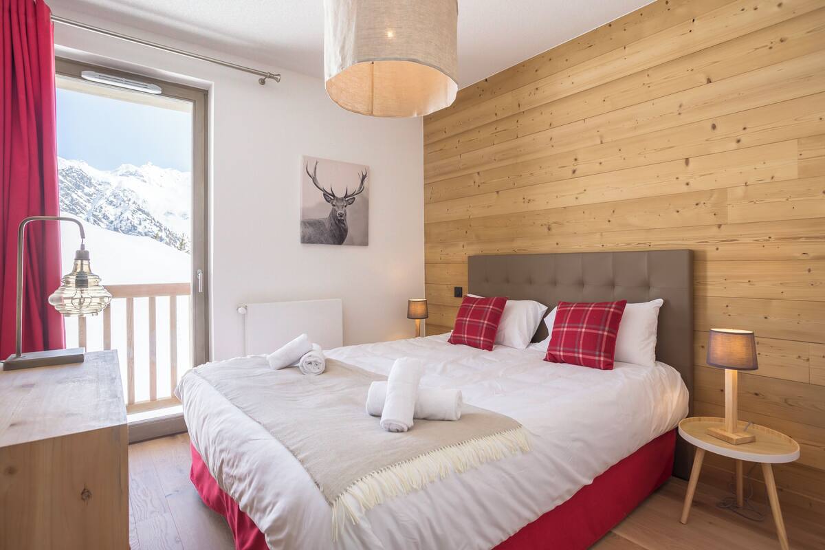 Une chambre d’hôtel avec un lit, un rideau rouge, un mur en bois, une fenêtre donnant sur la montagne et une décoration murale représentant une tête de cerf.