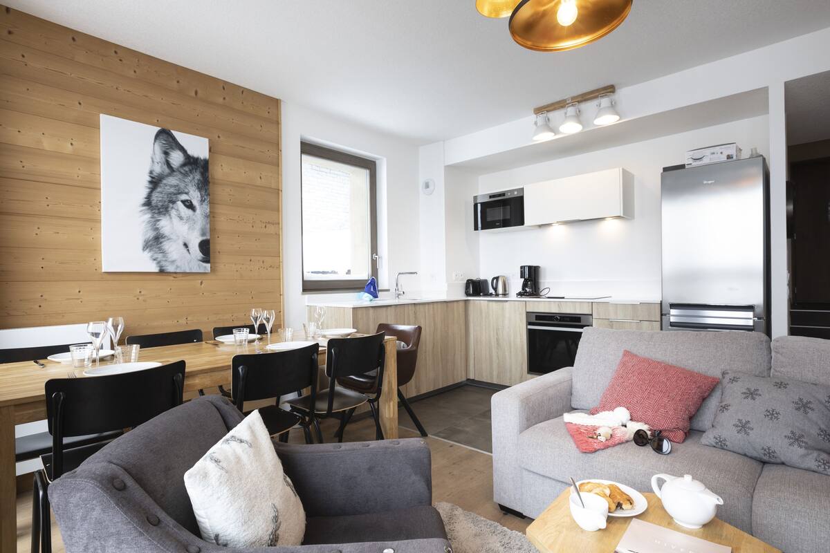 Un salon moderne avec un canapé gris, une table à manger pour quatre personnes et un coin cuisine équipé d’un four à micro-ondes et d’un réfrigérateur.