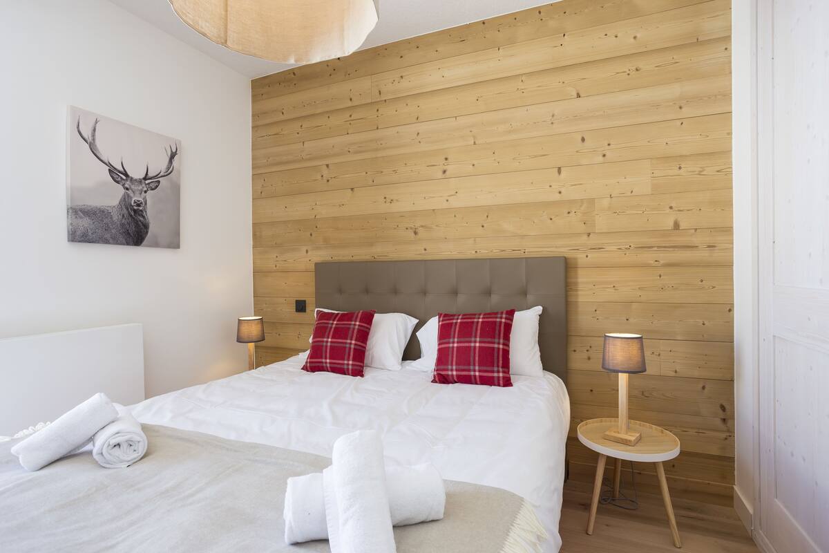Une chambre d’hôtel avec un mur en bois, un lit avec une literie blanche, des oreillers rouges et blancs, une petite table ronde en bois avec une lampe, et un tableau représentant un cerf, encadré, accroché au mur.